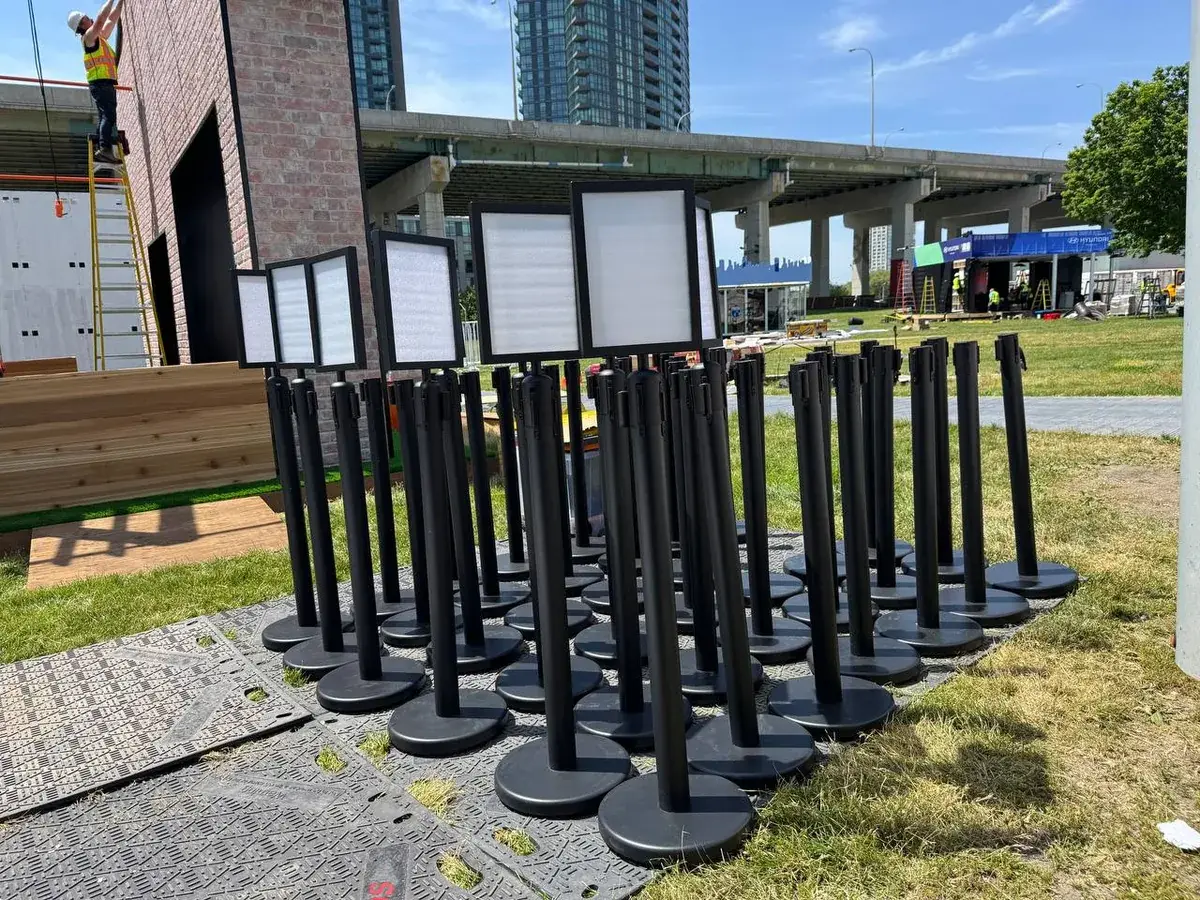 Black posts carrying printed sign frames, standing in a line on grass beside a timber boardwalk at an outdoor site.