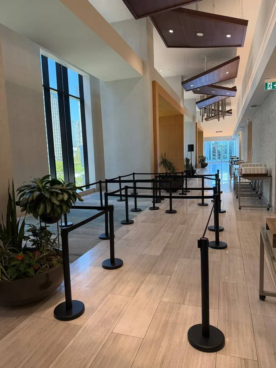 Black belt stanchions folded into a switchback across a wide interior with a wooden floor and planting along the wall.