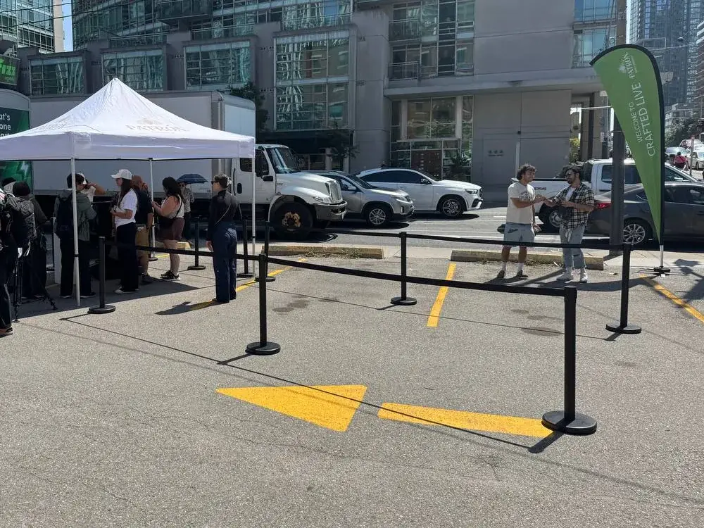 Black posts and belts closing off a section of street beside a pop-up canopy, with parked cars and a feather flag along the far edge.