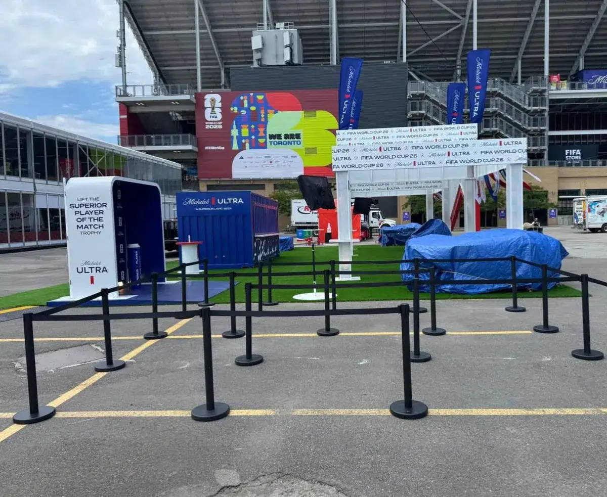Black belt stanchions marking out lanes on an asphalt forecourt at a stadium, with tournament banners and a strip of artificial turf behind them.