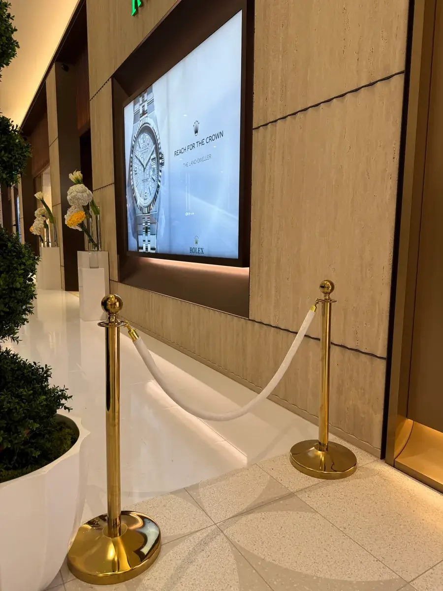 A white rope slung between two brass posts in a stone-lined lobby, planted troughs along the left and an illuminated display panel in the wall beyond.
